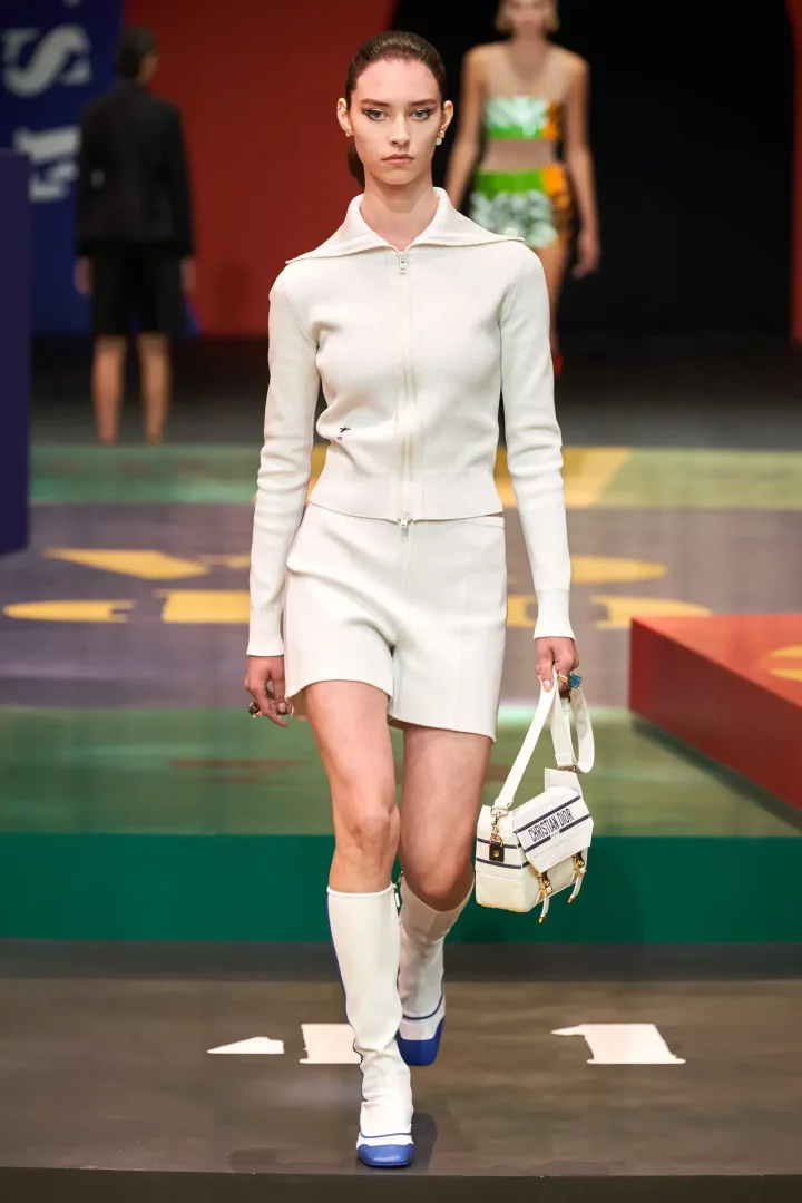 Podyumda yürüyen bir model, Christian Dior'un 2022 İlkbahar hazır giyim koleksiyonundan, beyaz yakalı fermuarlı uzun kollu üst ve şort takımını, Christian Dior yazılı beyaz çantayı ve mavi detaylı beyaz çizme kombinini sergiliyor.