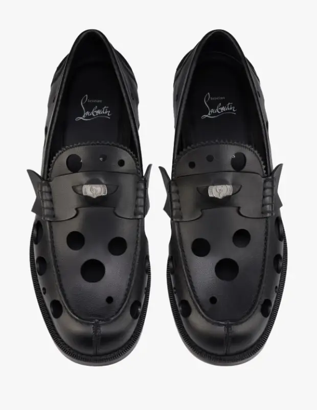 Christian Louboutin sonbahar-kış koleksiyonundan, üst kısmında karakteristik dairesel delikler ve gümüş renkli metal bir toka detayı bulunan, parlak siyah deriden yapılmış bir çift modern loafer ayakkabı, beyaz bir arka plan üzerinde dikkat çekiyor.