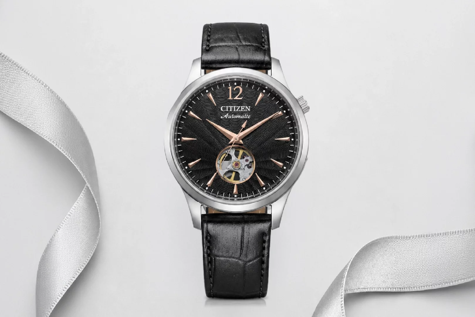 Beyaz bir zeminde, gümüş renkli kasası, rose gold ibreleri, açık mekanizma özelliği ve timsah desenli siyah deri kayışıyla dikkat çeken zarif bir Citizen Automatic klasik kol saati, Babalar Günü için ideal bir hediye olarak gümüş bir kurdele ile sunuluyor.