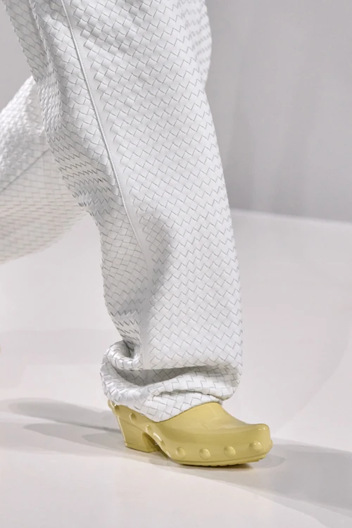 Beyaz örgü dokulu pantolon giyen bir modelin ayağında, önünde yuvarlak kabartma detayları bulunan pastel sarı renkte kalın tabanlı bir clog sandalet, 2026 İlkbahar-Yaz sezonunun öne çıkan sandal modelleri arasındaki yerini zanaat odaklı estetiğiyle sergiliyor.