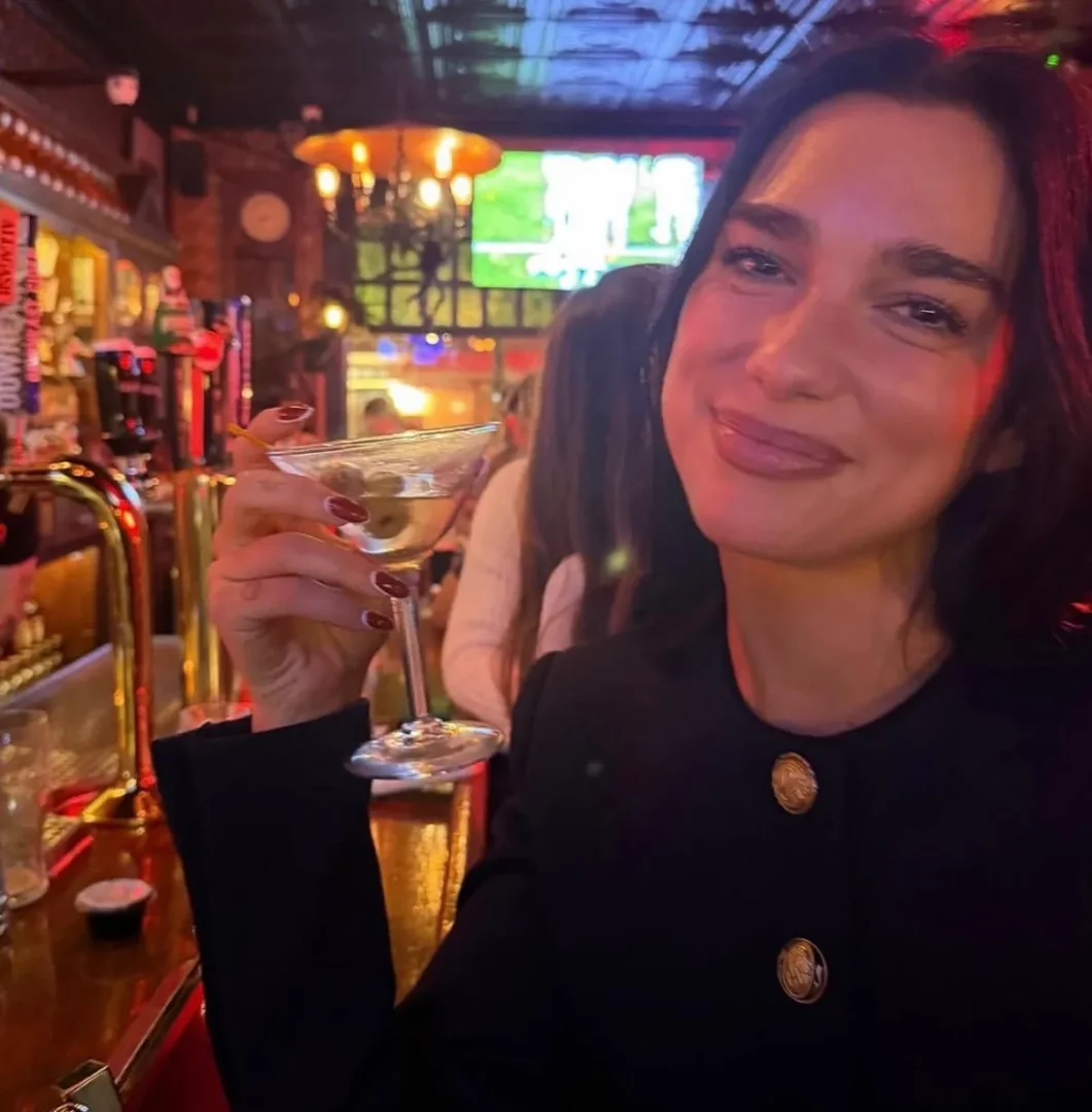 Koyu renk, düğmeli bir ceket giyen, kırmızı ojeli tırnaklarıyla zeytinli bir kokteyl kadehini tutan, loş ışıklı ve sıcak bir bistro bar ortamında, arka planda diğer misafirler ve parlak ekranlar eşliğinde içtenlikle gülümseyen bir kadın.