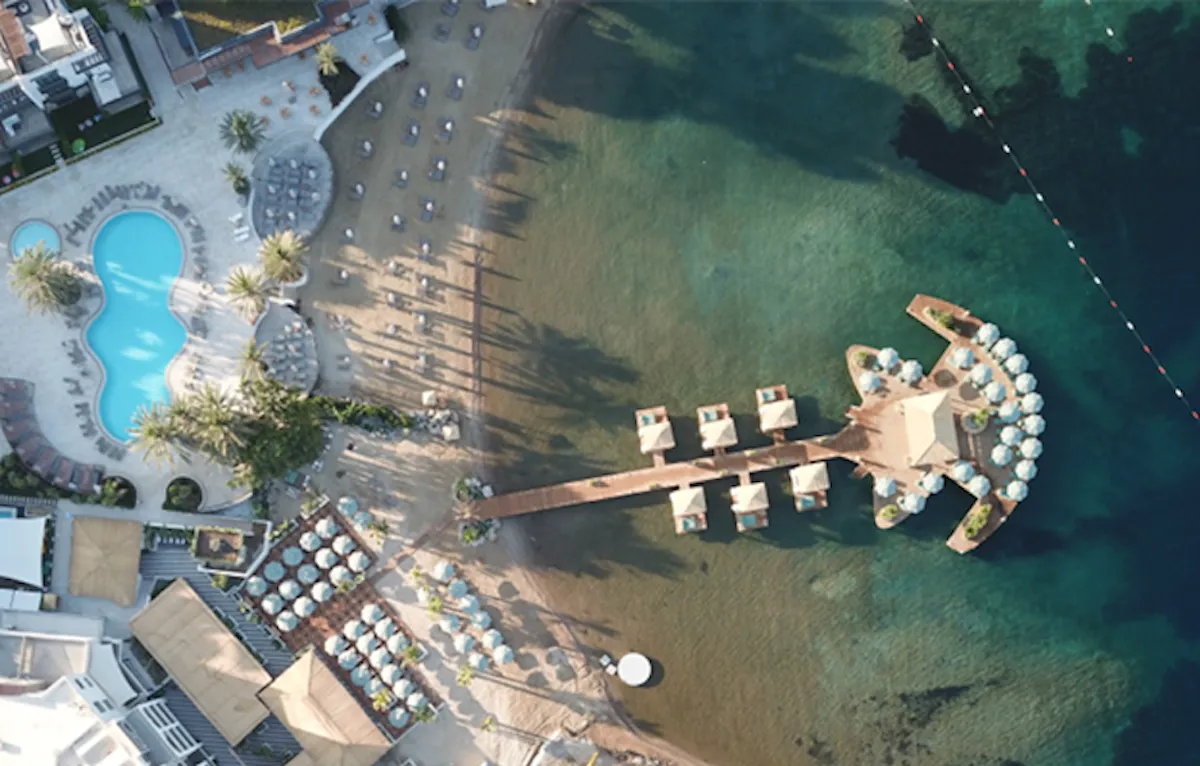 Kuşbakışı görünümde, Bodrum Yalıkavak'ta lüks bir sahil tesisine ait berrak turkuaz deniziyle buluşan kumlu plaj, havuz alanı, ahşap iskele üzerindeki şık su üstü bungalovlar ve iskelenin ucundaki çok sayıda şemsiye ile donatılmış geniş, gitara benzer özel bir platform dikkat çekiyor.