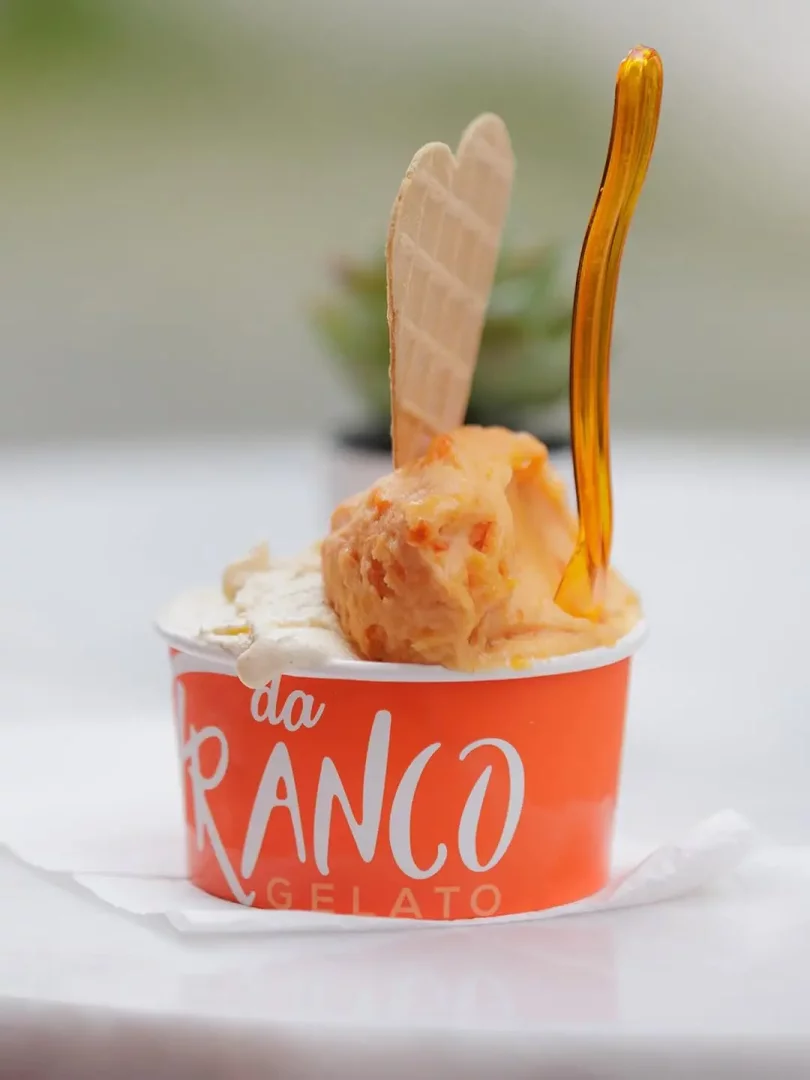 Parlak turuncu renkli da Franco Gelato bardağında, üzerinde kalp şeklinde bir waffle bisküvisi ve turuncu şeffaf bir kaşık bulunan, açık ve turuncu renkli iki top gelato, beyaz bir zemin üzerinde hafifçe bulanık yeşil bitki fonuyla iştah açıcı görünüyor.