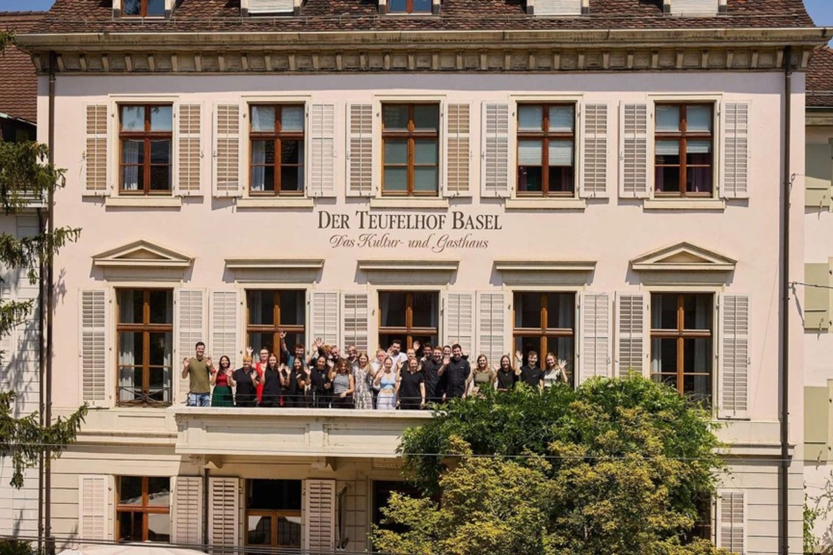 Soluk pembe cepheli, pencerelerinde panjurlar bulunan tarihi Der Teufelhof Basel Kültür ve Misafirhanesi'nin dış cephesinde, orta kattaki balkonda neşeyle el sallayan kalabalık bir grup insan yer alıyor.