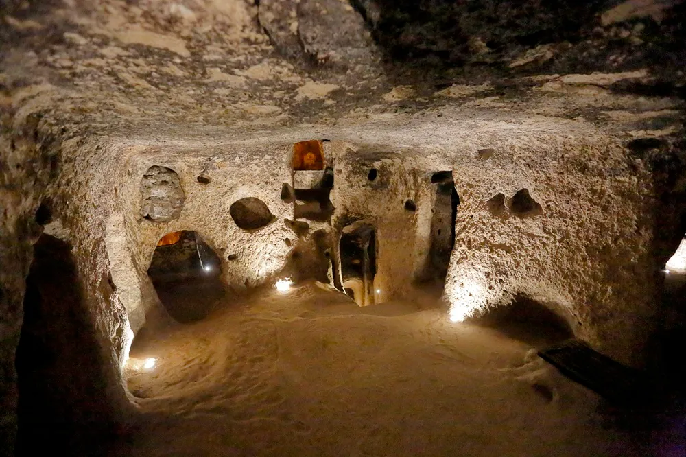 Kapadokya'daki Derinkuyu Yeraltı Şehri'nin bu aydınlatılmış bölümünde, kaba oyulmuş taş duvarları ve tavanıyla dikkat çeken geniş bir geçitte, çeşitli odacıklara ve koridorlara açılan kemerli girişler ile zemindeki spot ışıkları mekanın tarihi derinliğini ve gizemli dokusunu vurguluyor.
