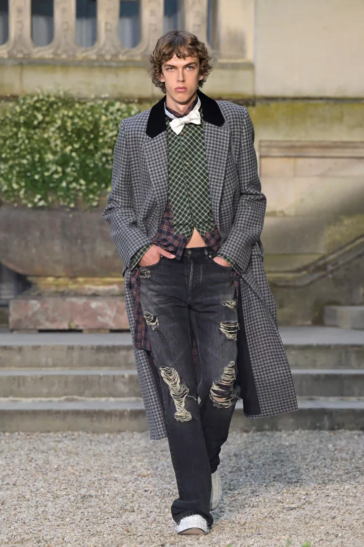 Dior 2027 İlkbahar-Yaz Erkek Koleksiyonu defilesinde, markanın klasik erkek giyim kodlarını beklenmedik bir şekilde yeniden yorumladığı kazayağı desenli uzun palto, yeşil baklava desenli yelek, kareli gömlek ve yırtık siyah jean pantolon giyen bir erkek model tarihi bir bahçede yürüyor.