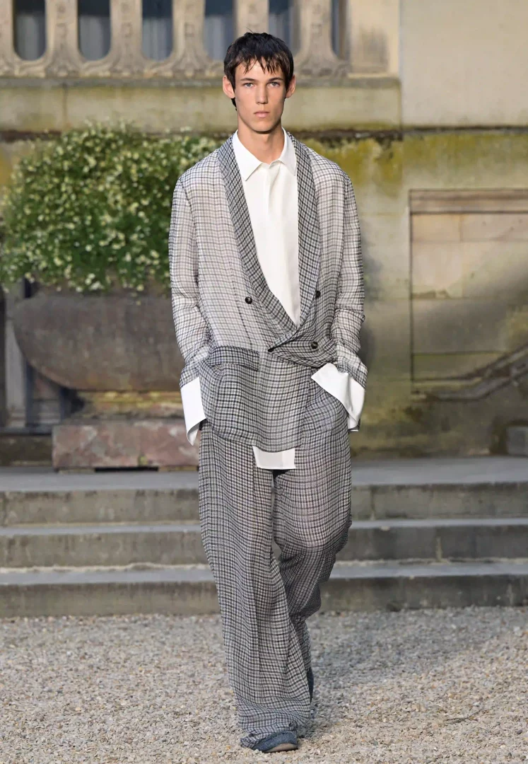 Dior'un 2027 İlkbahar-Yaz Erkek koleksiyonundan bir görünüm sergileyen genç model, klasik mimarinin önünde rahat kesimli, gri-beyaz kazayağı desenli kruvaze bir ceket ve bol paça pantolon giyerek markanın geçmişi günümüzle harmanlayan yeniden yorumlama temasını yansıtıyor.