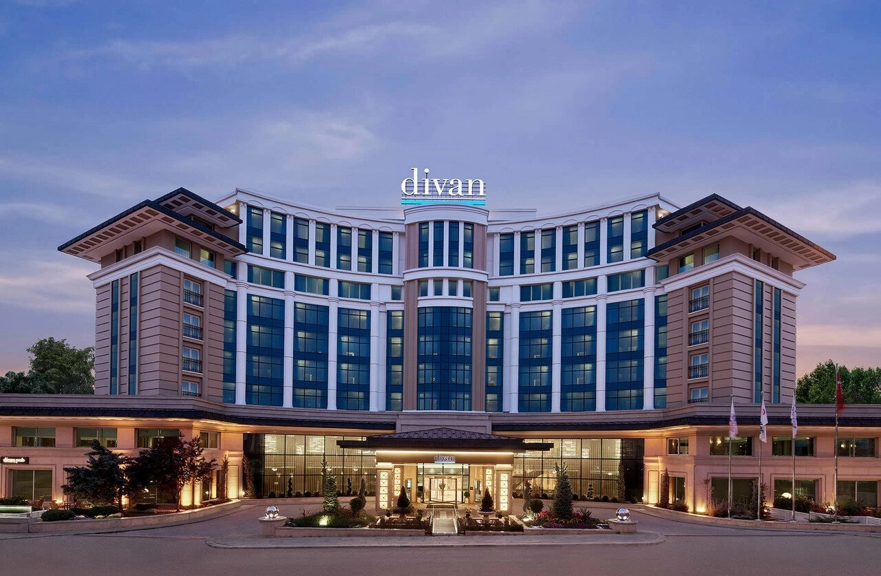 Divan Ankara'nın kavisli ve modern dış cephesi, bej ve beyaz tonlardaki mimarisiyle, akşamüstü ışıklarında parlayan mavi cam pencereleri ve şık girişindeki düzenli peyzajıyla, butik düğünler ve seçkin davetler için Ankara'da öne çıkan zarif bir düğün mekânını gözler önüne seriyor.