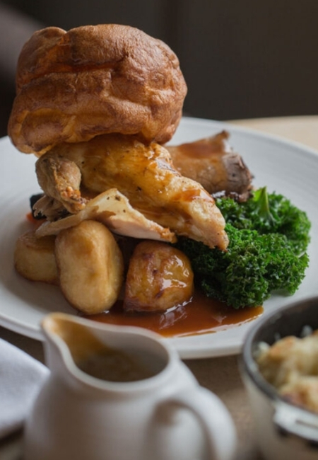 Yakın çekimde, kızarmış patates ve kıvırcık lahana ile zengin sosla sunulan, üzerinde büyük bir Yorkshire pudingi ve kızarmış tavuk parçaları olan bir tabak, Wimbledon Village'ın klasik pub'larında bulunan doyurucu İngiliz mutfağını temsil ediyor.