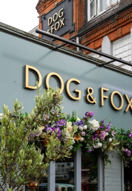 Wimbledon Village'da maç sonrası buluşma noktası olarak öne çıkan The Dog & Fox pub'ının, ön cephesindeki altın renkli DOG & FOX tabelası ve girişini çevreleyen mor, beyaz ve yeşil tonlardaki canlı çiçek süslemeleriyle Wimbledon ritüelini yansıtan dış cephesi.