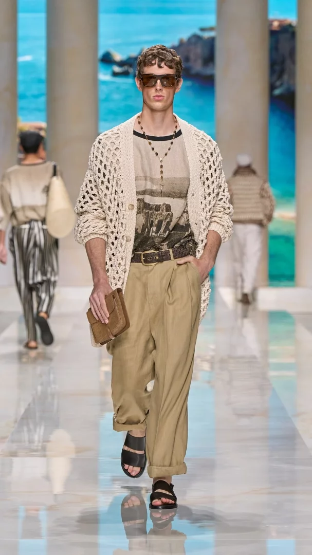 Milano Erkek Moda Haftası İlkbahar-Yaz 2027 Dolce&Gabbana defilesinde, model örgülü krem hırka, grafik desenli açık renk tişört, bol kesim keten pantolon ve sandaletlerle Akdeniz'in sıcak yaz atmosferini yansıtan şık bir görünümle podyumda yürüyor.