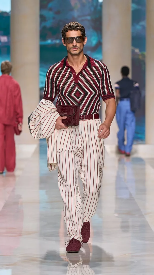 Milano Erkek Moda Haftası İlkbahar-Yaz 2027 defilesinde, Dolce&Gabbana'nın Akdeniz yaz esintili koleksiyonundan, bordo ve krem rengi geometrik desenli örme polo tişörtü, dikey çizgili pantolonu ve uyumlu ceketini taşıyan bir erkek model, belinde kroko desenli çanta ve bordo bağcıklı ayakkabılarıyla gözlükleri takılı podyumda yürüyor.