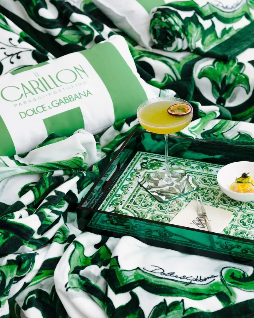 Dolce & Gabbana'nın Le Carillon Portofino ile yaptığı beach club iş birliğinde, Maiolica Verde temalı yemyeşil ve beyaz yaprak desenli havlu örtüler üzerinde, markanın logosunu taşıyan yastık ve çarkıfelek meyveli kokteyl ile şık bir tepsi yer alıyor, 2026 yazının lüks tatil deneyimini temsil ediyor.