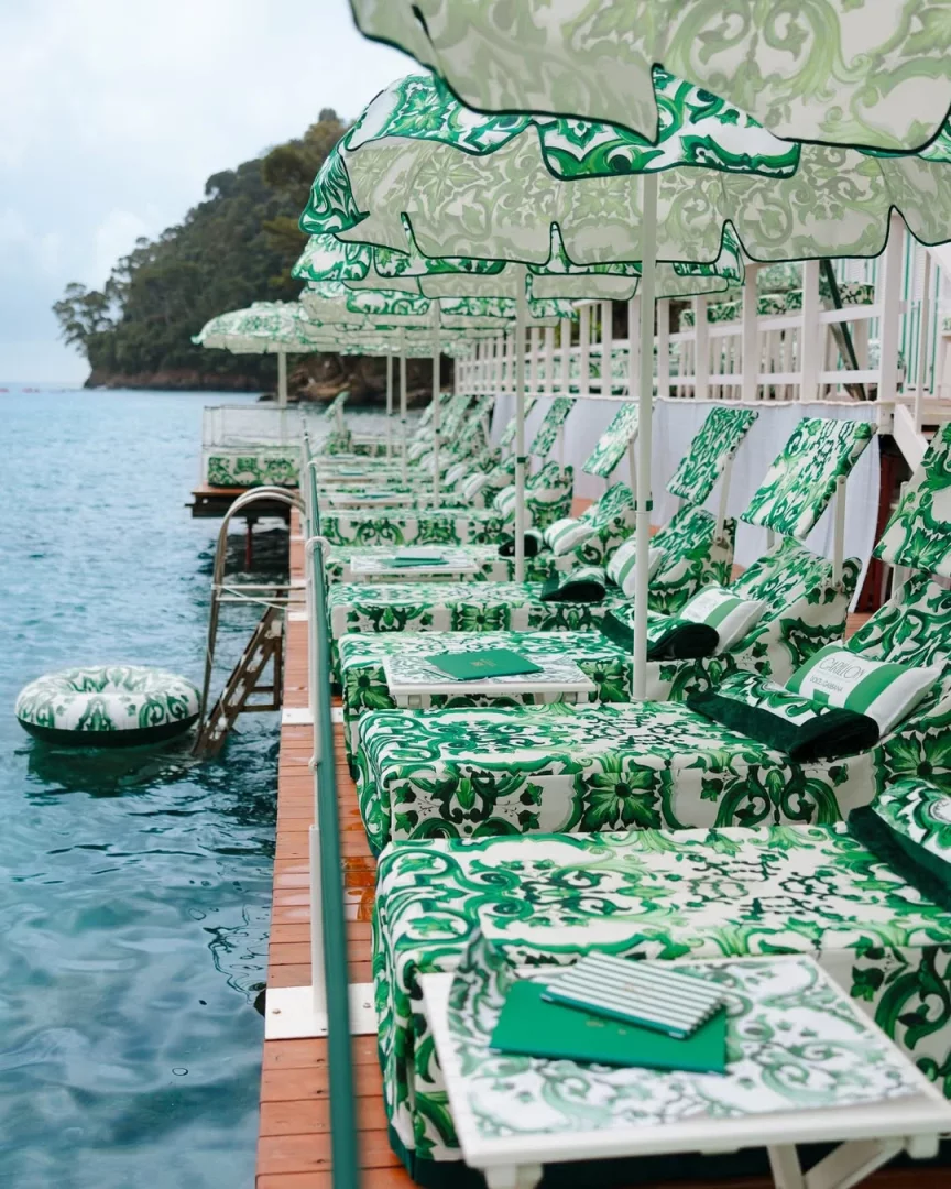 Portofino'daki Le Carillon beach club'ın Maiolica Verde temalı özel tasarımında, yemyeşil tepelerin ve turkuaz denizin kıyısında, Dolce & Gabbana'nın karakteristik yeşil-beyaz seramik desenleriyle bezenmiş şemsiyeler ve şezlonglar bir sıra halinde uzanıyor, markanın 2026 yaz tatili deneyimine şık bir dokunuş katıyor.