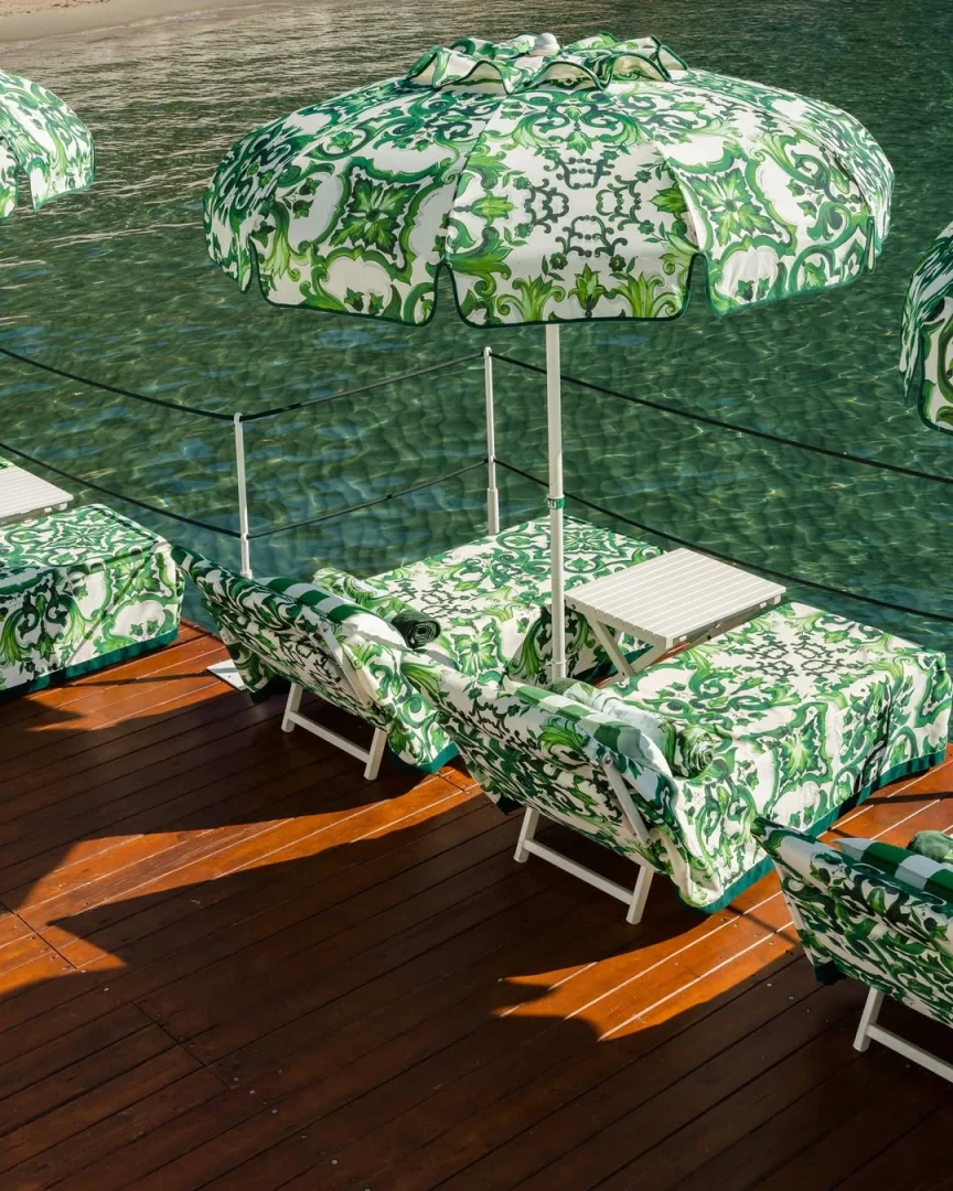 Dolce & Gabbana'nın 2026 yazının en çok konuşulan beach club iş birliklerinden biri olan Portofino'daki Le Carillon Beach Club'ı devralması kapsamında, markanın Maiolica Verde temalı yeşil ve beyaz seramik desenleriyle kaplı şemsiyeler ve şezlonglar, Akdeniz'in turkuaz suları üzerinde lüks bir tatil deneyimini gözler önüne seriyor.