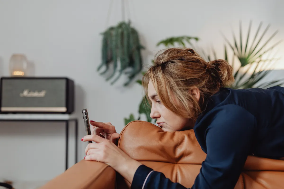 Kahverengi deri kanepede uzanmış, eliyle telefon ekranını kaydıran, yüzünde yorgun veya sıkılmış bir ifade olan bir kadın, modern çağın getirdiği dijital bağımlılık ve sürekli yapay uyarılmanın yol açtığı dopamin dengesizliği veya dopamin direnci sorununa işaret ediyor.