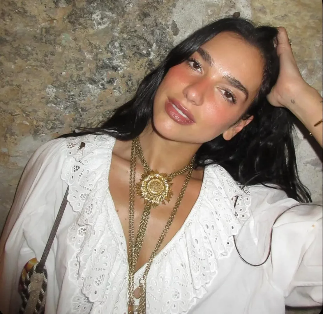 Dua Lipa, zarif beyaz dantelli fırfırlı bluzu, katmanlı altın renkli kolyeleri ve özellikle dikkat çeken büyük ayçiçeği ucuyla göz kamaştırırken, koyu dalgalı saçlarını eliyle geriye atıp hafifçe gülümseyerek kameraya bakıyor.