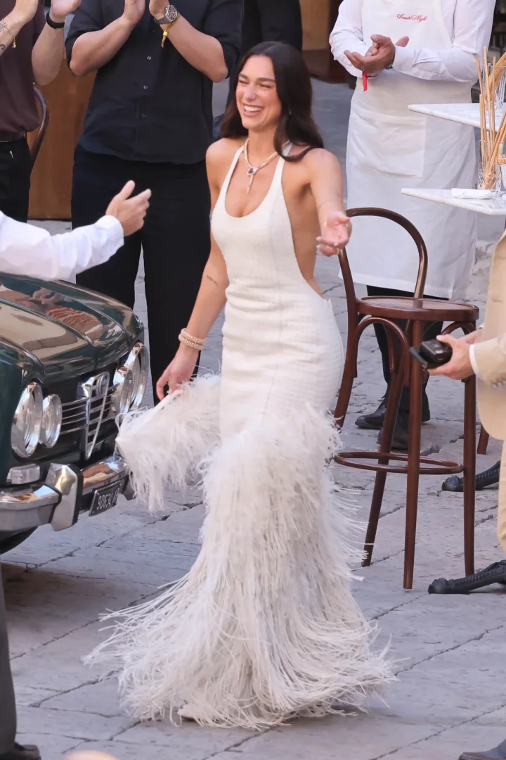 Dua Lipa, sırtı dekolteli ve etek kısmı gösterişli tüy detaylarıyla bezeli bembeyaz elbisesiyle neşeyle gülümsüyor, etrafındaki kutlama atmosferinde modern ve cesur düğün stilini sergileyen moda dolu anlar yaşanıyor.