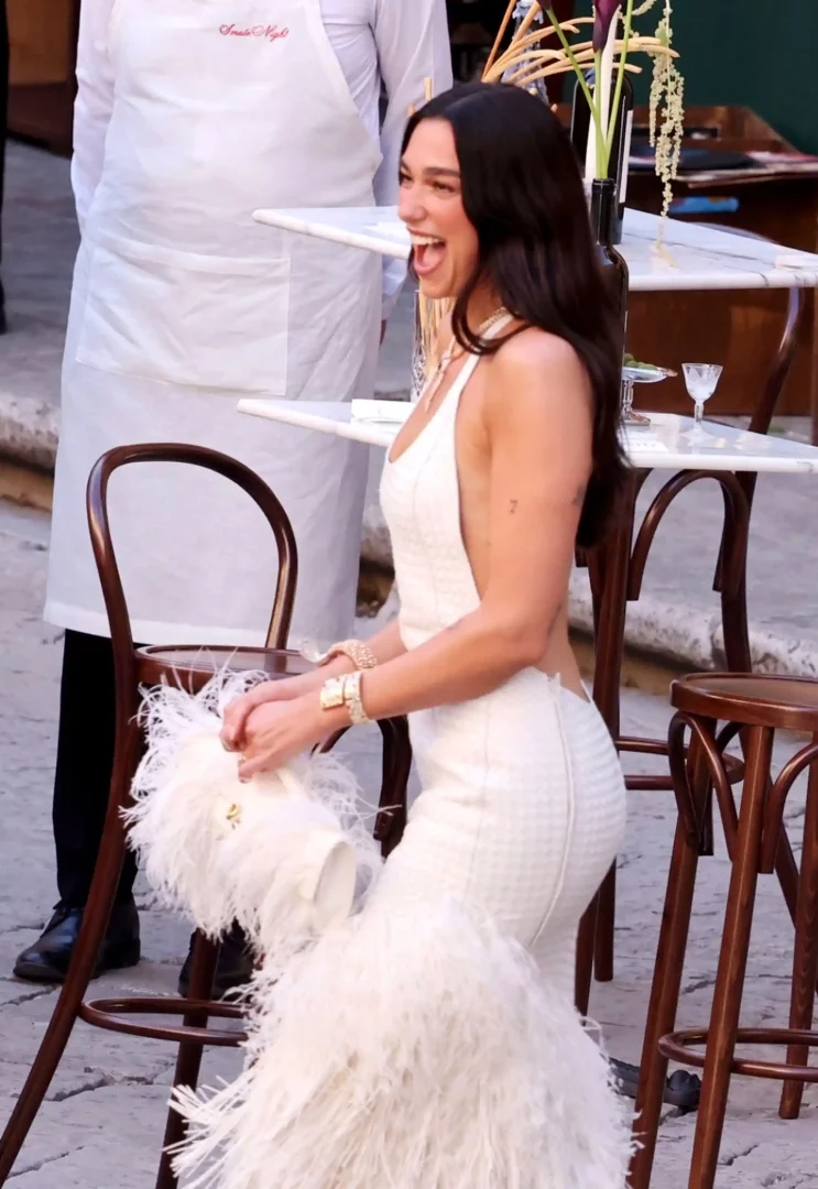 Dua Lipa, etekleri ve zarif çantası bembeyaz tüylerle süslü, sırtı açık, dokulu şık bir elbise içinde, dış mekân masalarının yanında genişçe gülümserken, moda dünyasının yakından takip ettiği düğün kutlamalarından birine ait keyifli bir anı yansıtıyor.