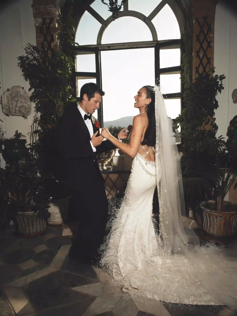 Sicilya'daki düğün kutlamalarında, beyaz sırtı açık, etekleri tüylü, işlemeli uzun bir Chanel Haute Couture gelinlikle dans eden Dua Lipa ile siyah smokinli Callum Turner, büyük kemerli pencereden görünen dağ manzarası eşliğinde neşeyle gülümsüyor.