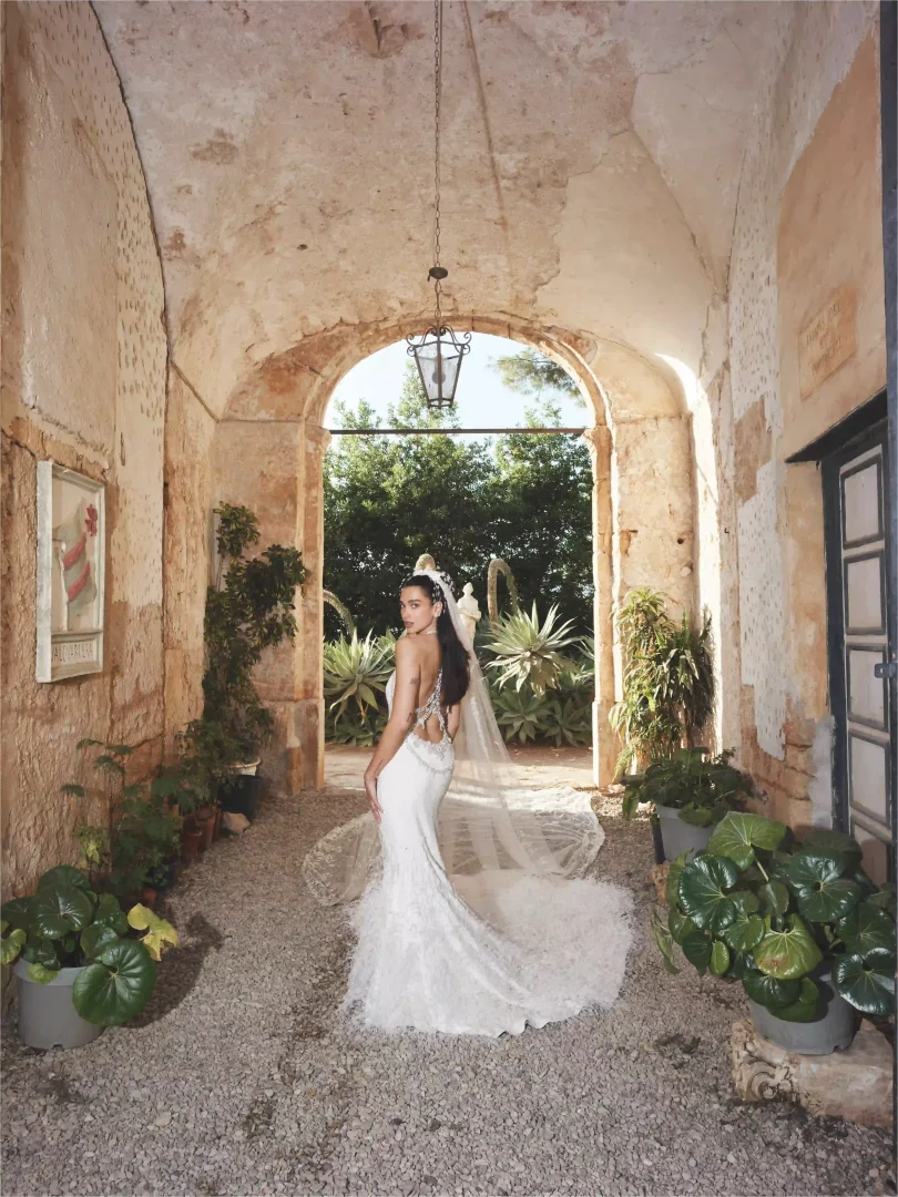 Dua Lipa, sırtı boncuk işlemeli ve uzun kuyruklu Chanel Haute Couture gelinliğiyle, rustik taş kemerli bir geçitte durup arkasına dönerek poz verirken, gelinliğin zarif işçiliği ve Sicilya'nın doğal atmosferi zamansız bir düğün görünümü sunuyor.