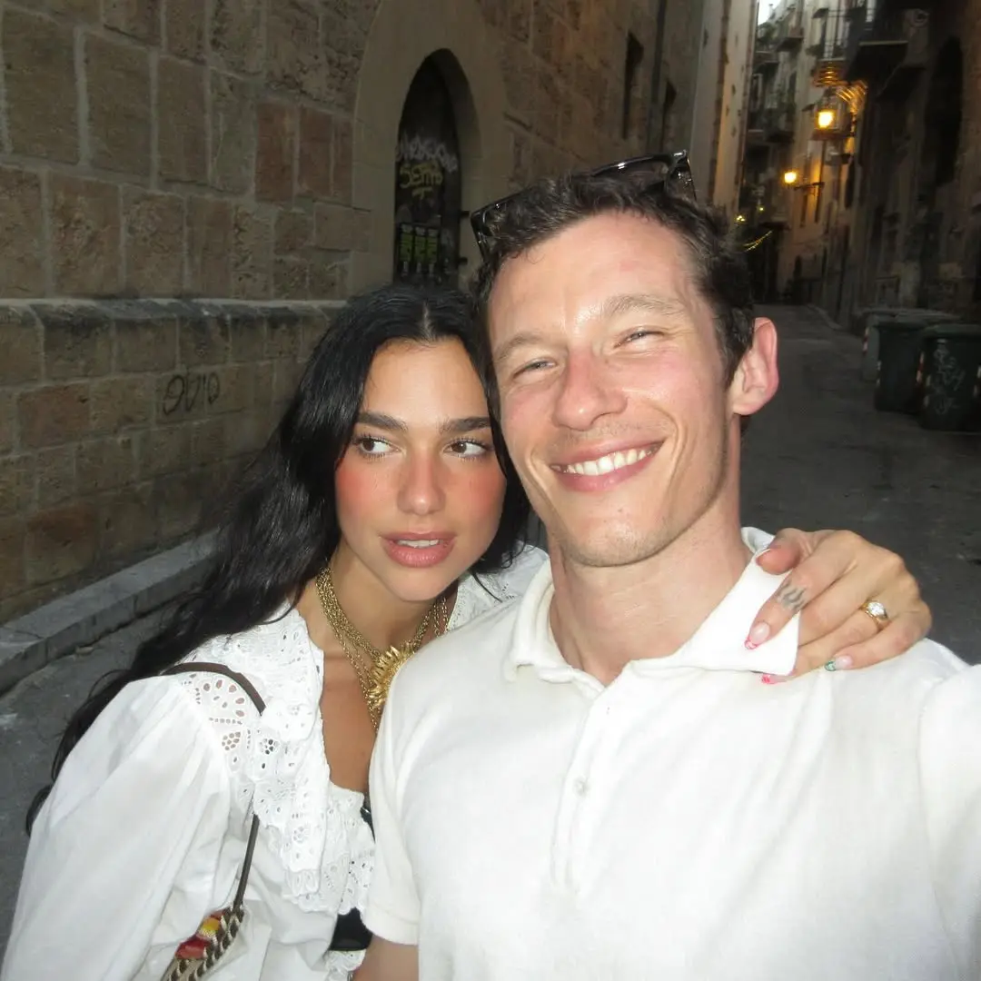 Dar bir sokakta gülümseyerek selfie çeken Dua Lipa ve sevgilisi Callum Turner, 2026-2027 nişan yüzüğü trendlerine ilham veren, kalın sarı altın bantlı ve yuvarlak kesim pırlantalı yüzüğünü sergiliyor.