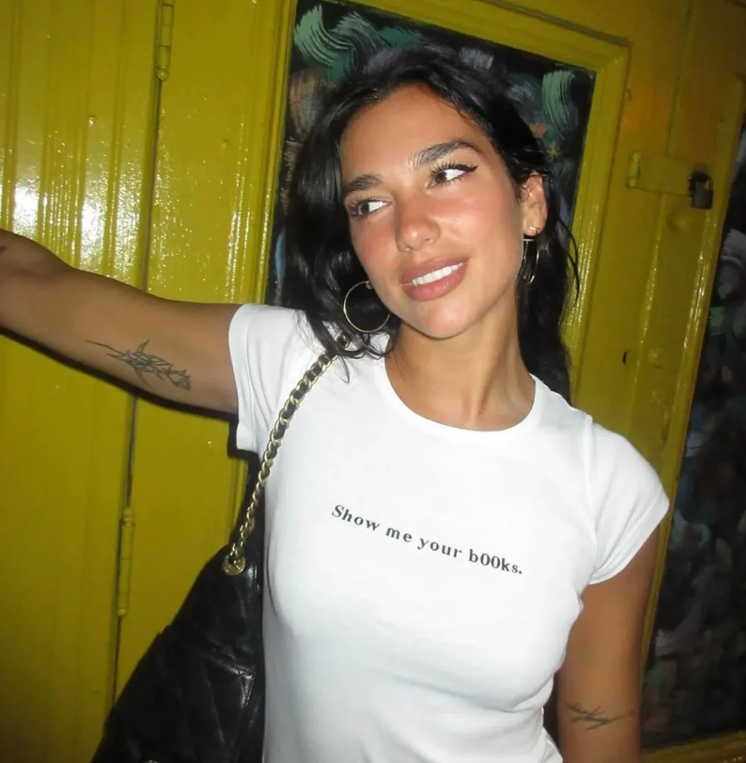 Dua Lipa, parlak sarı bir arka planın önünde, Show me your books yazılı beyaz bir tişört, büyük altın halka küpeler ve zincir askılı siyah bir çanta ile hafifçe gülümseyerek yan tarafa bakarken, kolundaki dövmesi de belirgin.