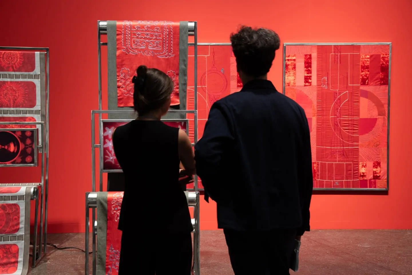 Kırmızı bir sanat galerisinde sırtları dönük iki ziyaretçi, canlı kırmızı tonlarda, beyaz ipliklerle karmaşık desenler işlenmiş tekstil sanat eserlerini inceliyorlar, bazıları raflarda sergilenirken bazıları parlak kırmızı duvara asılı.