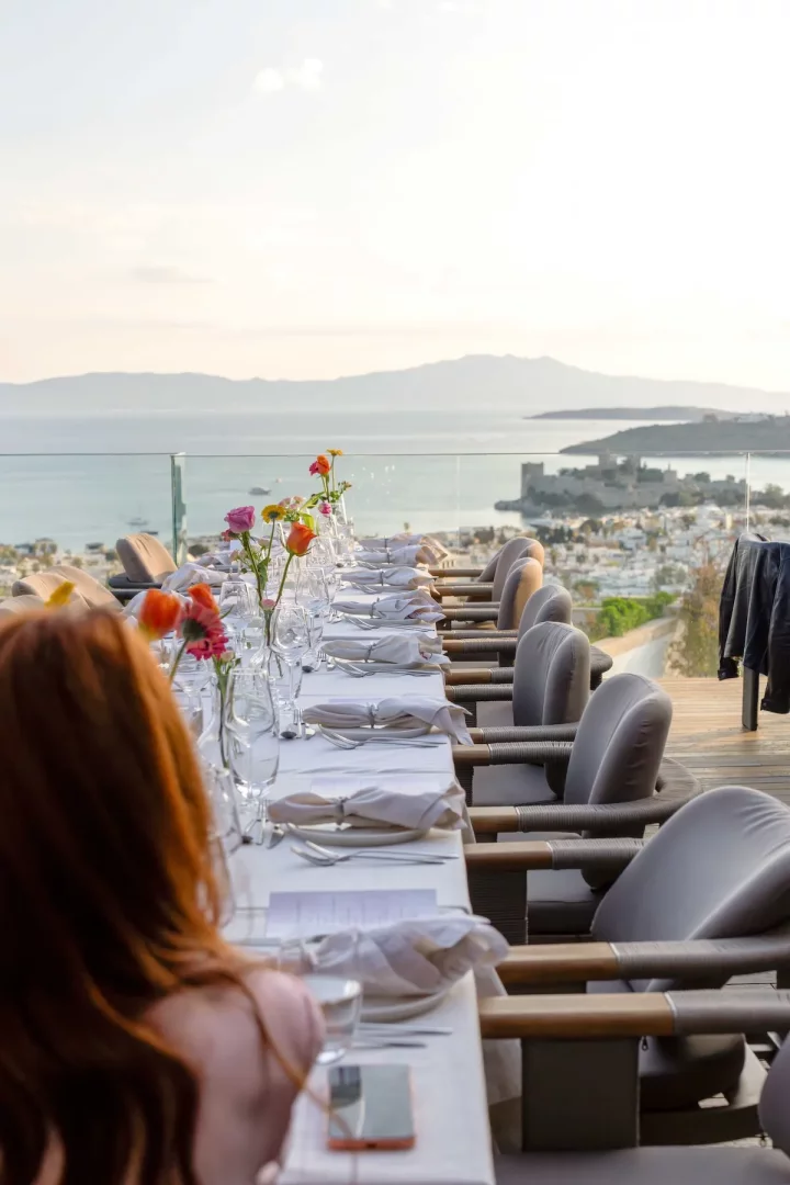 Bir restoran terasında, renkli çiçeklerle ve şık servis takımlarıyla donatılmış uzun bir masa, masmavi Ege Denizi, Bodrum Kalesi ve uzaktaki dağların büyüleyici manzarasına karşı gün batımı ışıklarında misafirlerini bekliyor gibi duruyor.