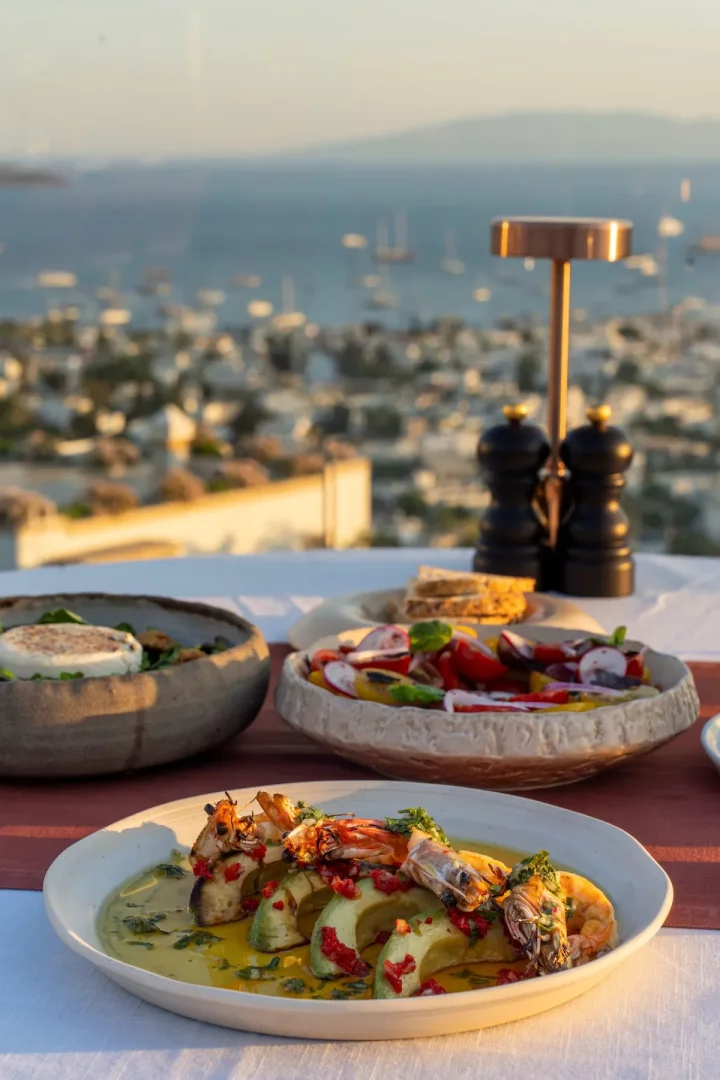 Bodrum'un eşsiz deniz manzarasına nazır bir terasta, gün batımı ışıkları altında özenle hazırlanmış ızgara karides ve avokado dilimleri içeren zarif bir ana yemek, yanında taze yeşillik ve domates salatalarıyla birlikte servis edilen Efil Restoran'ın keyifli atmosferini yansıtan bir görsel.