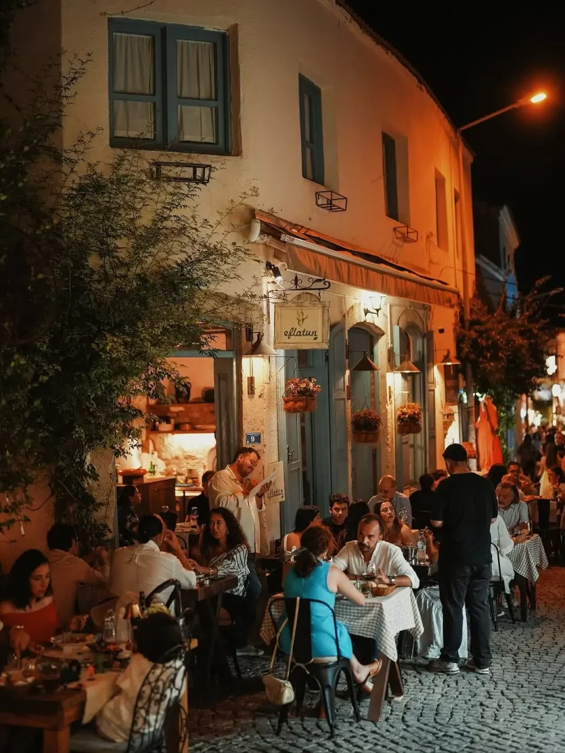 Alaçatı'da Eflatun restoranın önünde Arnavut kaldırımlı sokakta, sıcak ışıklarla aydınlatılmış beyaz cepheli binanın önünde, çiftlerin ve arkadaş gruplarının canlı bir akşam yemeği keyfi yaptığı kalabalık ve samimi bir açık hava mekanı.