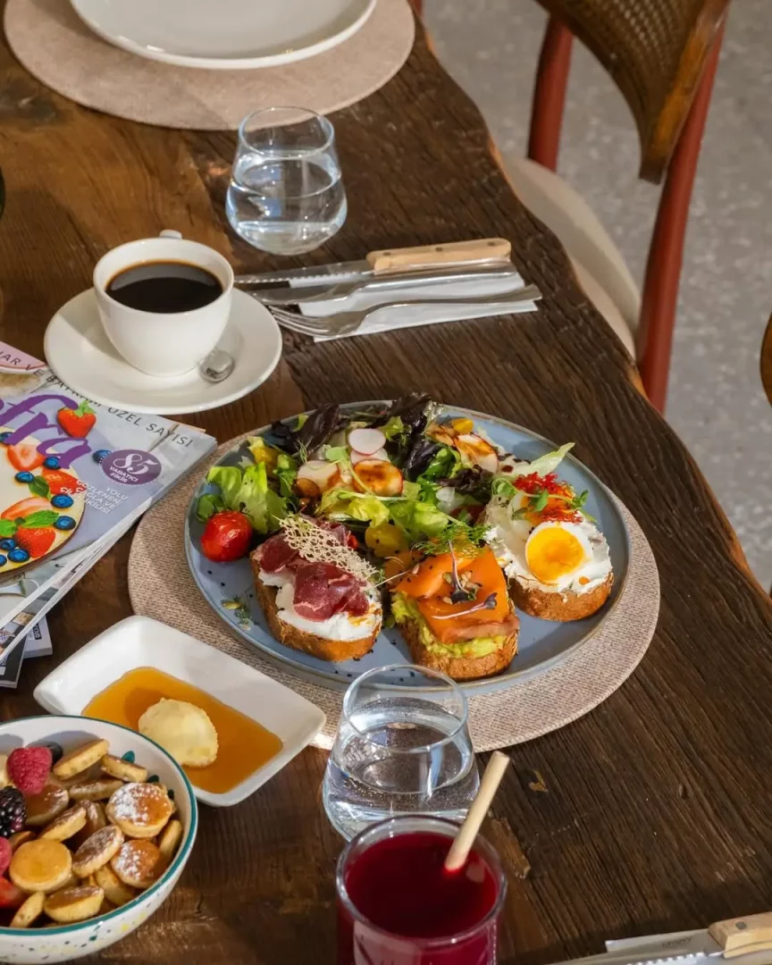 Rustik ahşap bir masa üzerinde, peynir, füme et, avokado-somon ve sahanda yumurta gibi çeşitli malzemelerle hazırlanmış bruschetta benzeri üç farklı kızarmış ekmek dilimi, taze yeşillik salatası, meyve ve pudra şekeri serpilmiş mini pankekler, bal, tereyağı, filtre kahve, su ve kırmızı meyve suyu içeren iştah açıcı bir kahvaltı veya brunch sofrası yer alıyor.