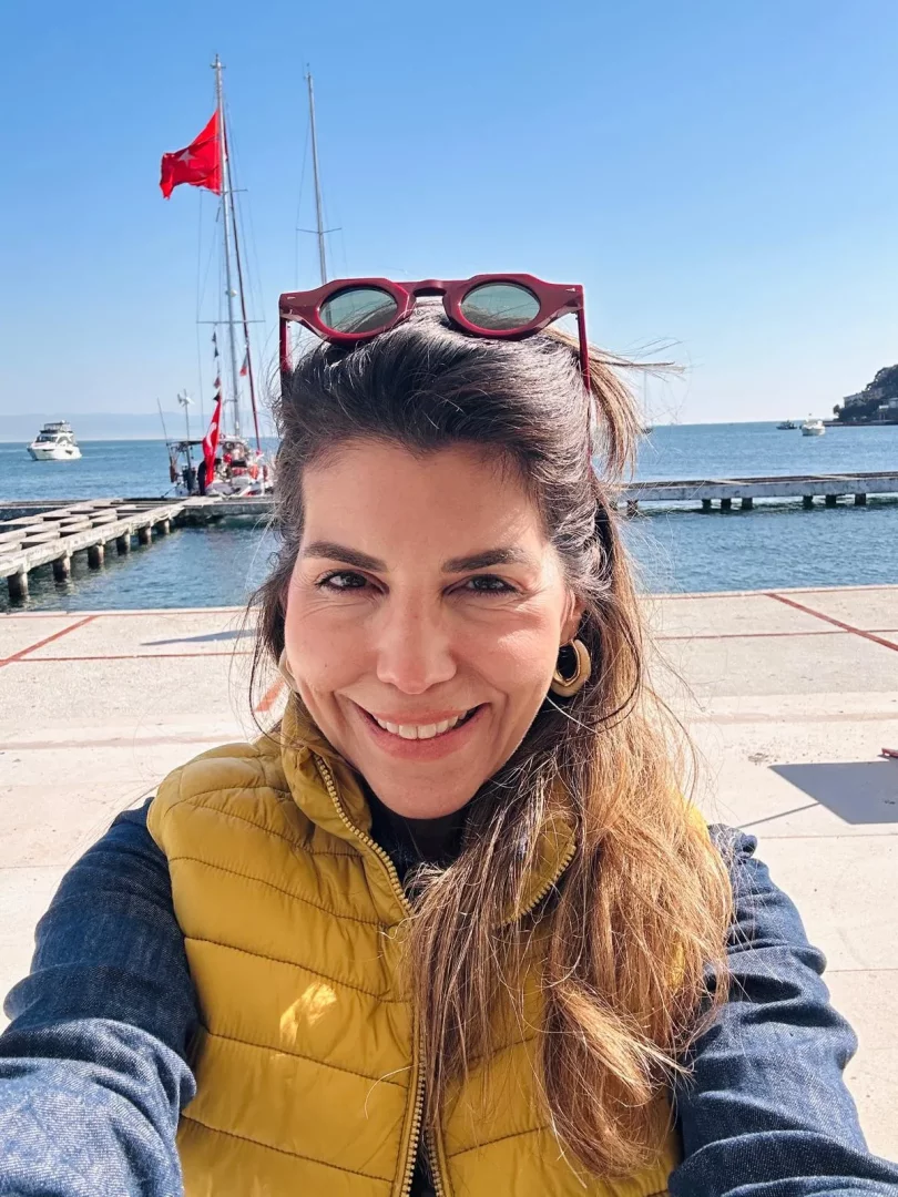 Elmon, saçlarının tepesine bordo güneş gözlüğü takmış, sarı şişme yeleği ve kot gömleğiyle gülümseyerek selfie çekiyor; arka planda masmavi gökyüzü, deniz üzerinde ahşap bir iskele ve Türk bayraklı bir yelkenli tekne görünüyor.