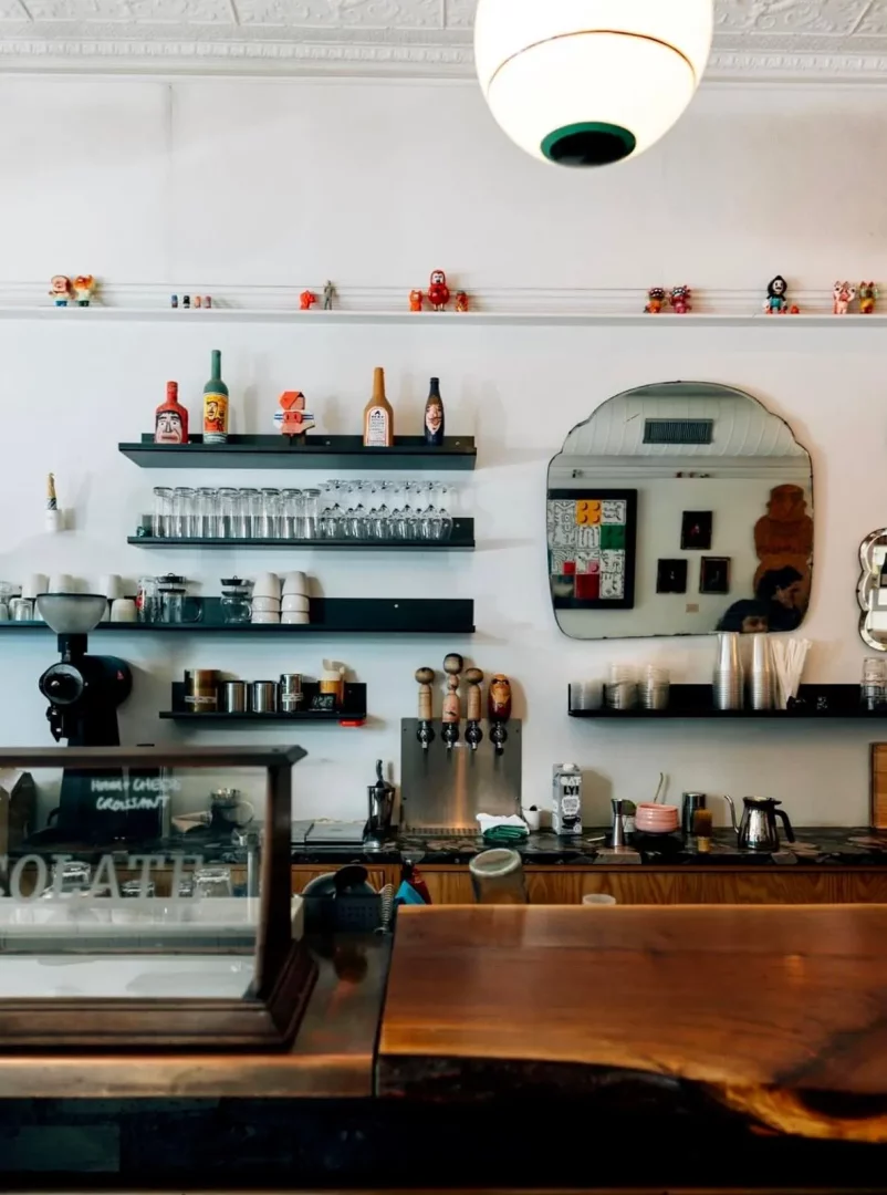 Beyaz duvarlı, ahşap tezgahlı ve modern sarkıt lambalı bir kafenin iç görünümü, üst raflarda renkli figürler, orta raflarda sıra sıra kadeh ve bardaklar, alt raflarda kahve makinesi ve musluklar, tüm bu düzenlemede sunulan çeşitli içecek ve yiyecek önerilerini sergiliyor.