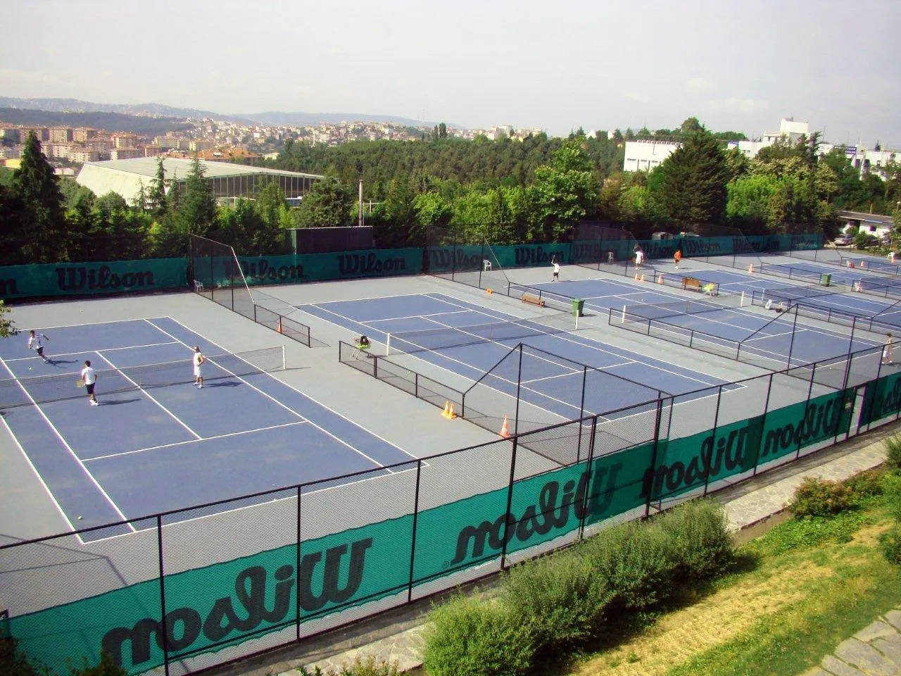 Enka Tenis Kulübü'nün kuşbakışı çekilmiş fotoğrafında, mavi ve gri zemine sahip çok sayıda tenis kortu görülüyor; kortlarda raketleriyle tenis oynayan ve dinlenen sporcular, Wilson logolu yeşil güvenlik çitleri ve arka planda ağaçlarla kaplı şehir manzarası dikkat çekiyor.