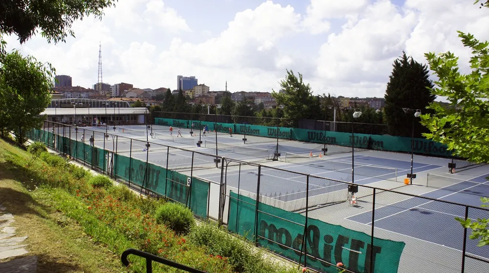 Enka Tenis Kulübü'nün çitlerle çevrili, yeşil Wilson pankartlarıyla süslenmiş gri ve mavi açık hava tenis kortlarında, birçok oyuncunun güneşli havada aktif bir şekilde tenis oynadığı geniş bir görüntü; arka planda şehir silüeti ve yeşil ağaçlar görülmektedir.