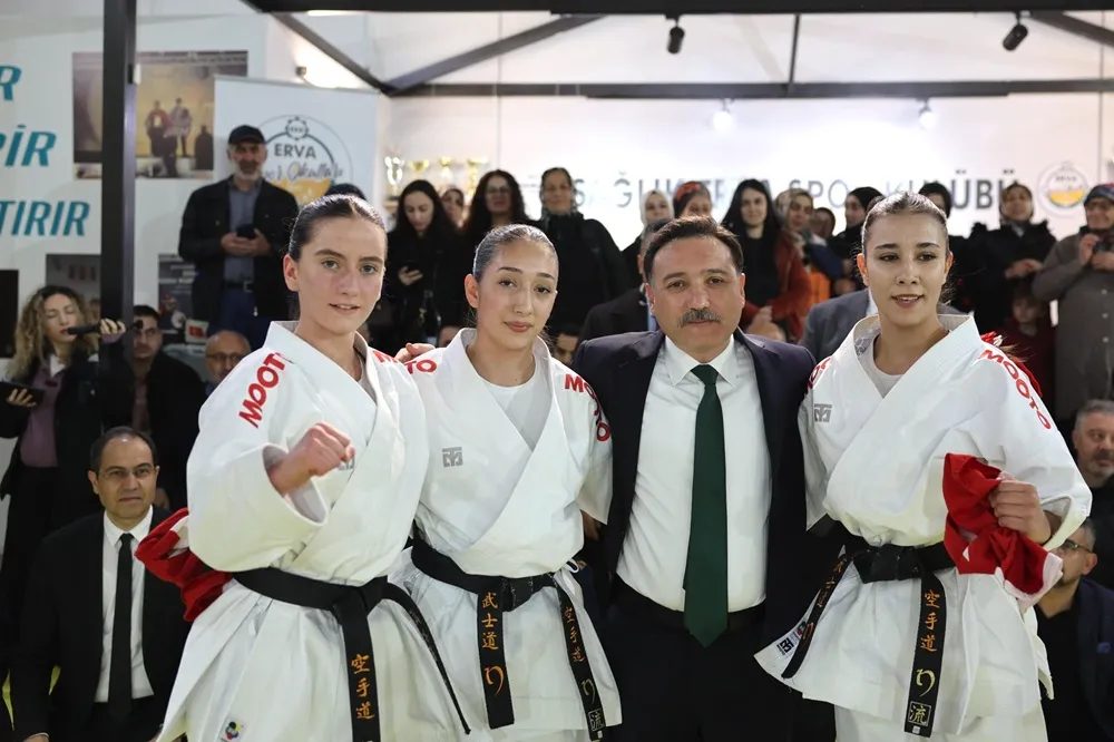 Erva Okulları'nın düzenlediği bir spor etkinliğinde, beyaz karate gisleri ve siyah kuşaklarıyla dikkat çeken üç kadın karateci ile koyu takım elbiseli bir erkek, kalabalık bir spor salonu ortamında, iki sporcunun elinde kırmızı bezler varken gururla poz veriyor.