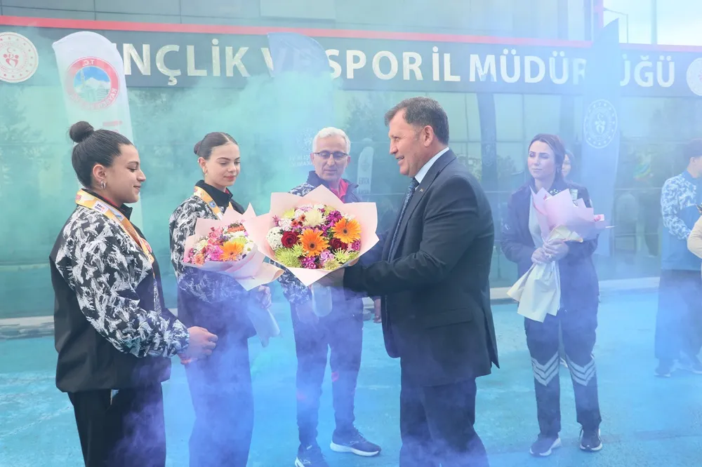 Gençlik ve Spor İl Müdürlüğü binasının önünde, mavi kutlama dumanları arasında, madalyalı iki tekvandocu Erva ve takım arkadaşı, takım elbiseli bir yetkiliden tebrik edilerek renkli çiçek buketleri alıyorlar.