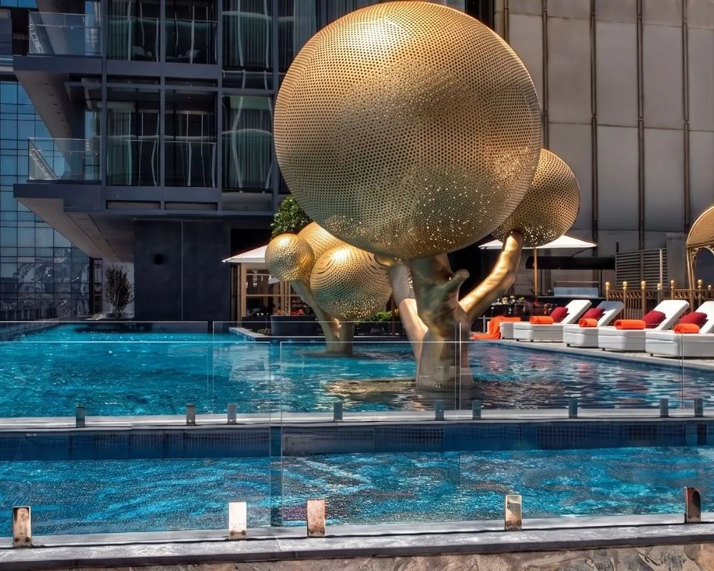 Fairmont Otel'in muhteşem dış mekan havuzu, berrak mavi suyun içinden yükselen parlak altın rengi, delikli küre biçimli modern sanat ağaç heykelleriyle dikkat çekiyor; havuzun etrafında kırmızı yastıklı ve turuncu havlulu rahat beyaz şezlonglar sıralanırken, şeffaf cam korkuluklar bu lüks ve modern atmosferi tamamlıyor.