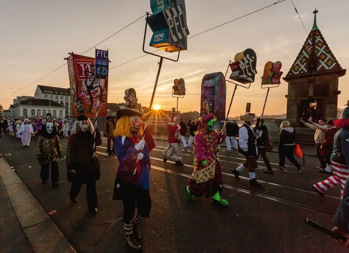 Basel gezilecek yerler rotasında Fasnacht karnavalı geçidinde maskeli katılımcılar, ışıklı fenerler ve gün batımı atmosferi.