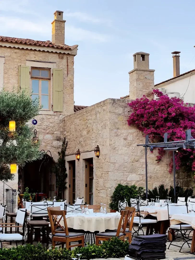 Alaçatı'nın karakteristik taş binaları arasında yer alan Fava adlı restoranın, beyaz masa örtülü ve ahşap sandalyeli zarif masaların özenle dizildiği, duvarlarında canlı mor begonvillerin sarktığı, zeytin ağaçlarından sarkan lambalarla aydınlatılan, davetkar ve huzurlu bir açık hava yemek alanı.