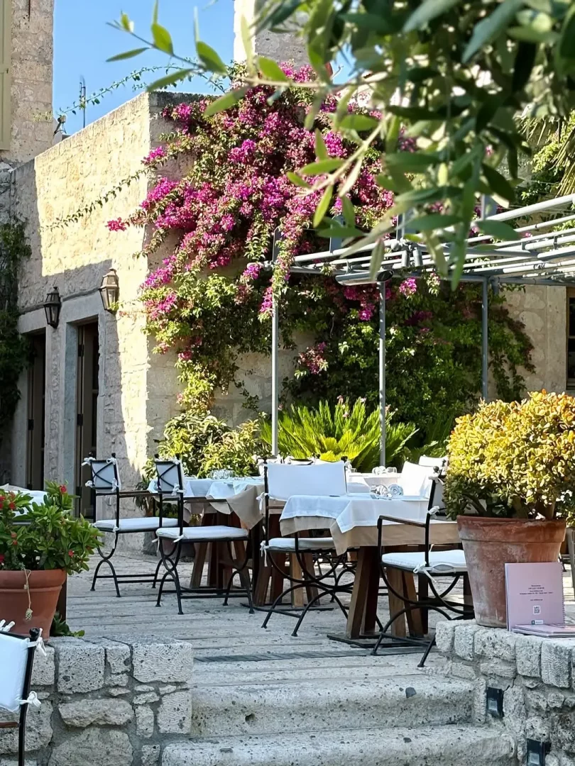 Alaçatı'nın taş evleriyle ünlü şirin sokaklarından birinde yer alan, canlı pembe begonvillerle sarılı taş duvarların önünde, beyaz örtülü masaları ve ferforje sandalyeleriyle misafirlerini bekleyen, tipik Ege mimarisine sahip açık hava restoran alanı.