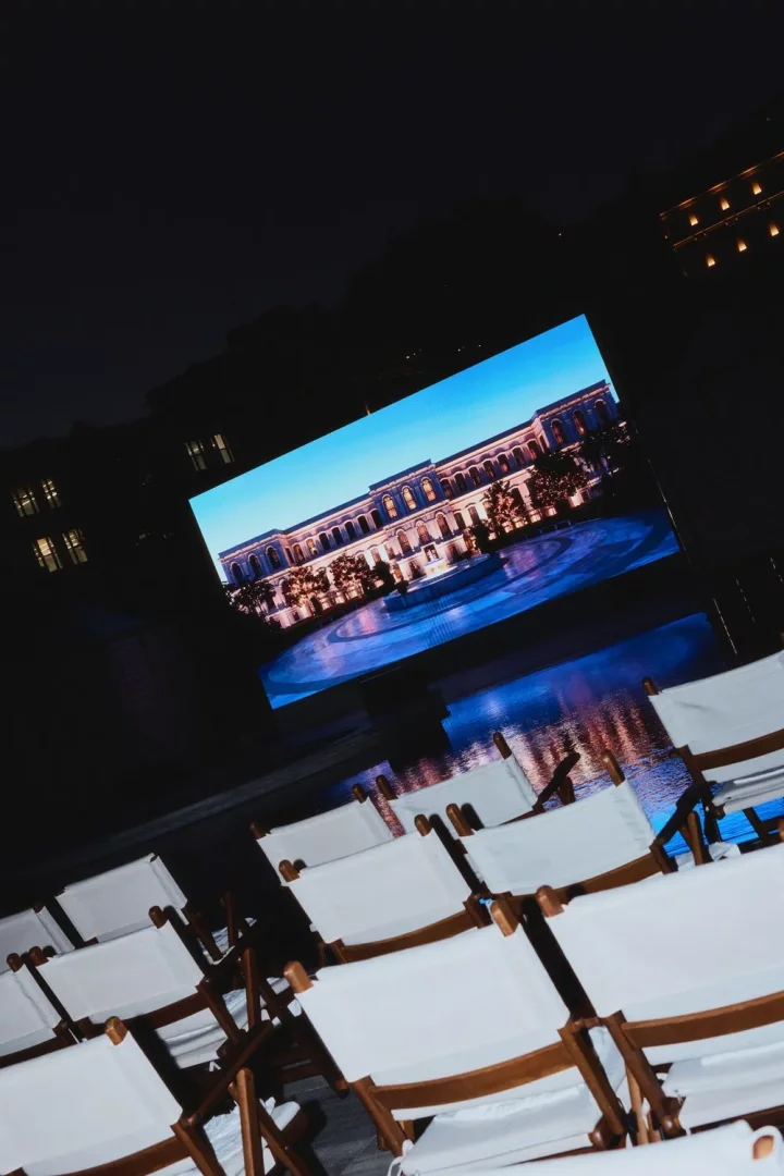 Gecenin karanlığında, lüks bir Four Seasons açık hava sineması etkinliğinde sıralanmış boş beyaz katlanır sandalyelerin önünde, ışıklandırılmış şık bir otel binasının görüntüsünü yansıtan büyük bir ekran görülüyor.
