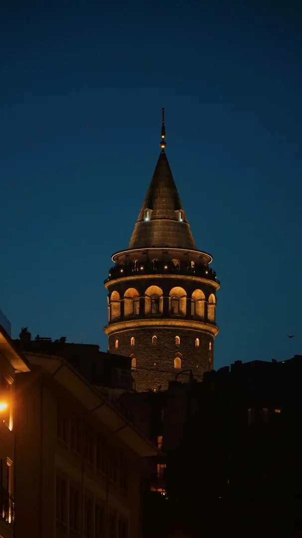Gece çökerken, koyu mavi gökyüzü altında parlayan, taş duvarları ve konik çatısıyla ışıl ışıl aydınlatılmış Galata Kulesi, gözlem güvertesindeki siluetleriyle tarihi bir müze olarak İstanbul'un eşsiz güzelliğini yansıtıyor.