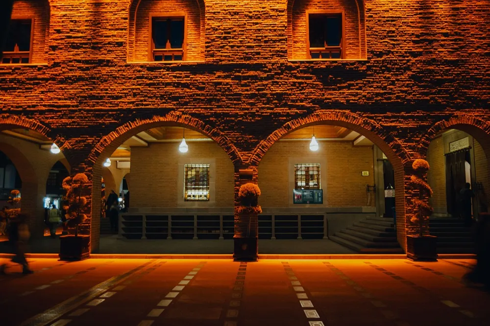 Gece karanlığında sıcak turuncu ışıklarla aydınlatılmış, tuğla duvarları ve kemerli girişi olan bu tarihi yapı, gece müzeleri etkinliği öncesinde ziyaretçileri ağırlamaya hazır, gizemli ve davetkar bir atmosfer sunuyor.