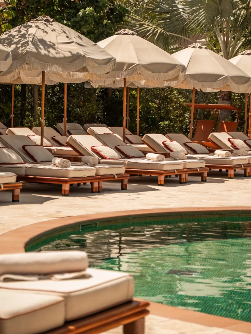 Bodrum'daki Gigi Beach'in havuz kenarında, bej minderli ve kırmızı yastıklı sıralanmış şezlongların üzerinde beyaz havluların durduğu, saçaklı büyük bej şemsiyelerin gölgelediği, yemyeşil bitki örtüsüyle çevrili, berrak yeşil mozaik kaplı havuzun sakin görüntüsü.