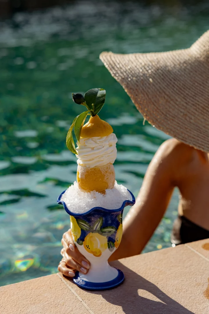 Havuz kenarında hasır şapkalı bir kadının elinde tuttuğu, özel tasarım limon desenli bir kupada buz içinde sunulan, limonlu dondurma topu, krema ve taze limonla zenginleştirilmiş ferahlatıcı bir Bodrum limonlu tatlı sunumu dikkat çekiyor.