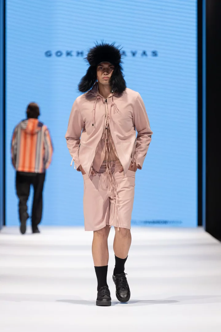 Gökhan Yavaş'ın podyumunda, siyah tüylü şapka, bağcıklı açık pembe kapüşonlu ceket ve uyumlu şort giyen bir erkek model, Haziran ayının öne çıkan moda haberlerindeki yeni tasarımları temsil eden bir duruş sergiliyor.