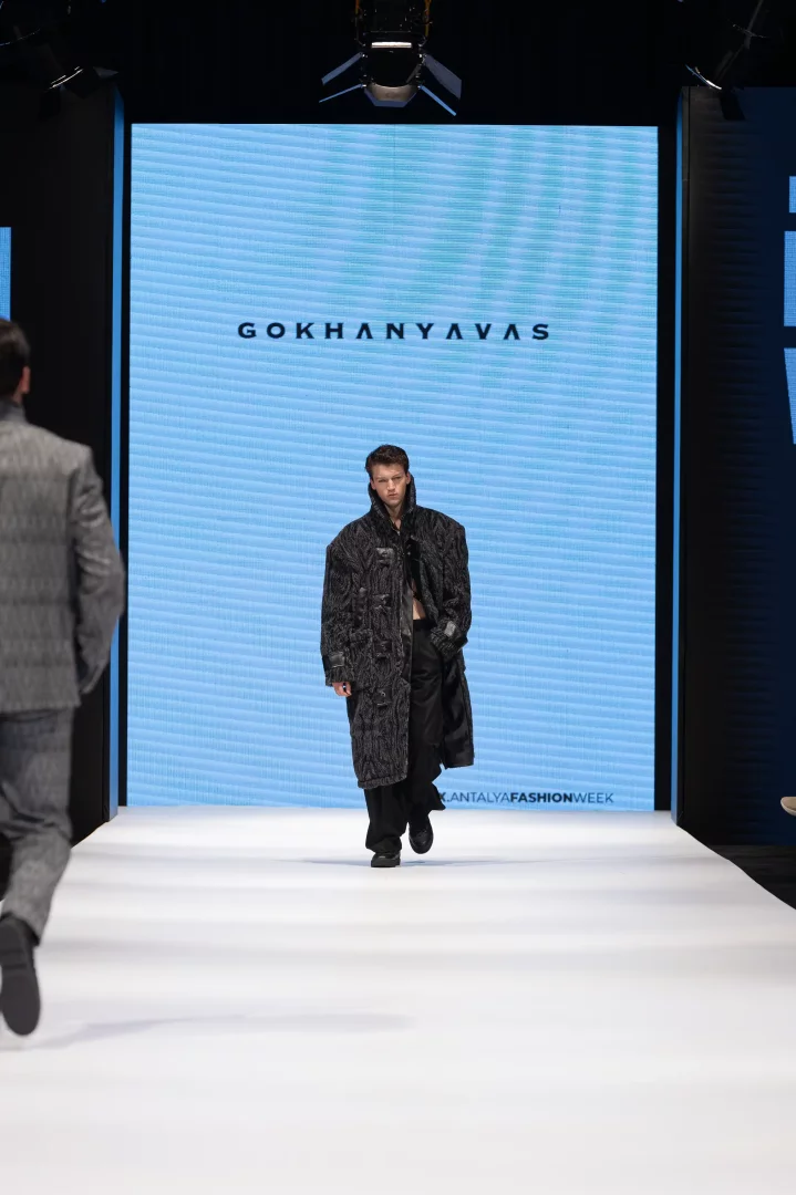 Antalya Fashion Week podyumunda, Gökhan Yavaş'ın sonbahar-kış koleksiyonundan, dokulu koyu gri oversized uzun bir palto ile geniş paça siyah pantolon giyen, modanın sınırlarını zorlayan çarpıcı bir yeni tasarım sergileyen erkek model.