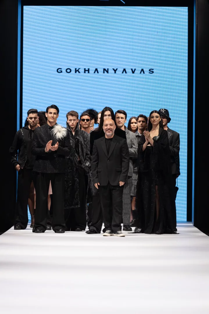 Moda dünyasındaki yeni tasarımları yansıtan bu karede, tasarımcı Gökhan Yavaş'ın podyumda modelleriyle birlikte durduğu görülüyor, arka plandaki ekranda GOKHANYAVAS yazısı belirirken modellerin giydiği gösterişli ve koyu renkli Sonbahar/Kış 2027 koleksiyonundan parçalar dikkat çekiyor.